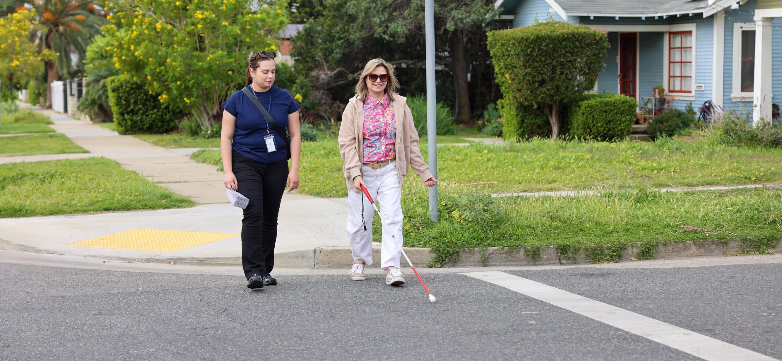 Woman crossing the street while using a cane. An orientation and mobility instructor walks beside her.
