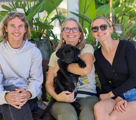 Three people sitting on a bench holding a black labrador puppy